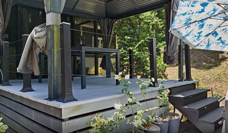 A small elevated deck with black railings and steps leading up to it, surrounded by potted plants and shaded by a large patterned umbrella on Roatan, Honduras.
