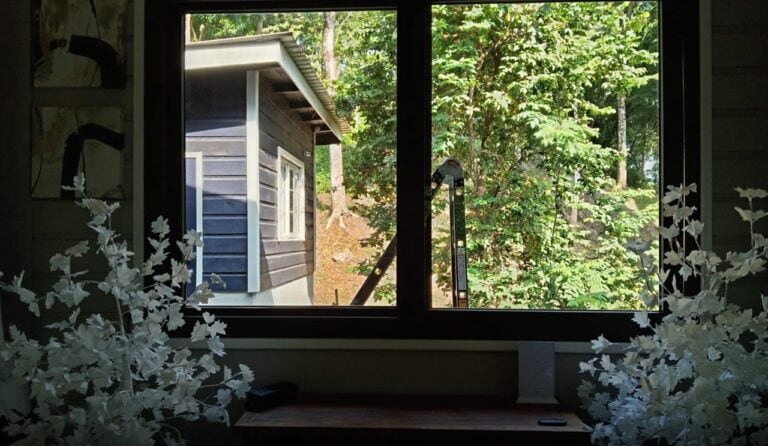 View through a window showing a small blue wooden house with white trim on a lush green forested hillside in Roatan, Bay Islands, Honduras.