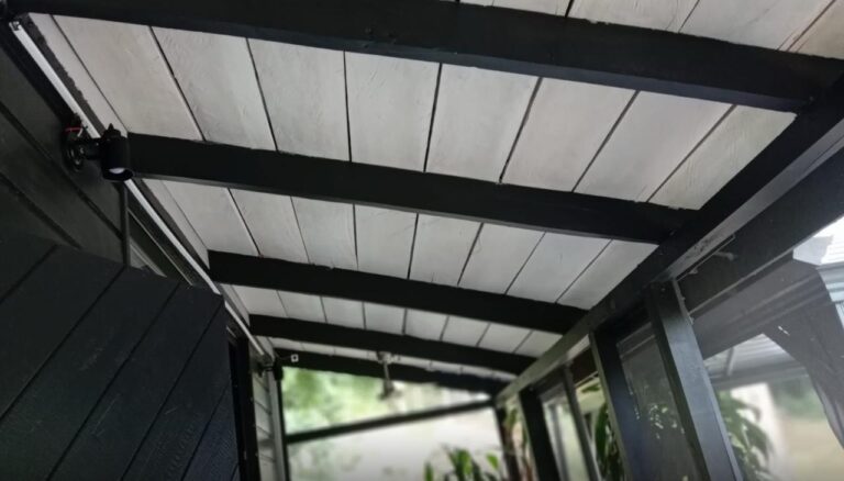 The image shows a close-up view of a covered outdoor patio area with black wooden beams and a white ceiling in Roatan, Honduras, featuring a black security camera mounted on the wall.