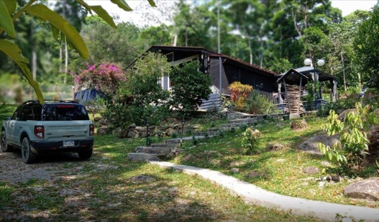 A small, rustic house with a sloped roof situated in a lush, green landscape in Roatan, Honduras, surrounded by tropical plants and trees while a white SUV is parked nearby.