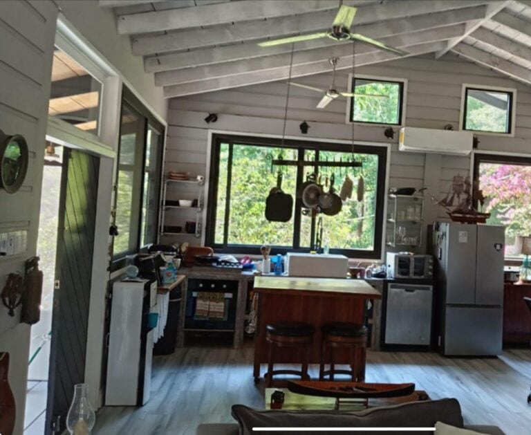 Inside view of a cozy wooden kitchen in Roatan, Honduras, featuring large windows with greenery outside, hanging pots and kitchen utensils, and a compact, functional layout.