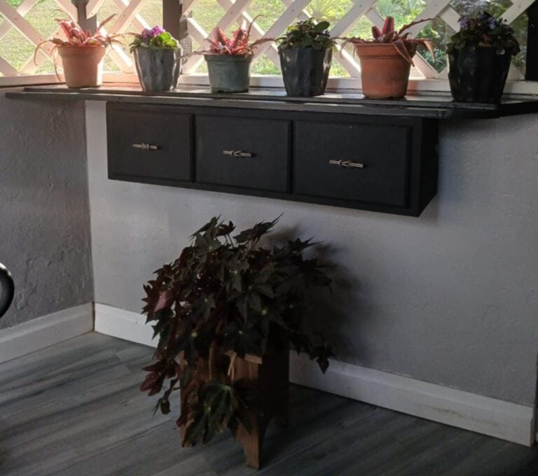 A black wall-mounted cabinet with three drawers adorned with metal handles, situated against a white textured wall near a corner of a room in Roatan, Honduras. Above the cabinet, a windowsill displays five potted plants with vibrant pink and green foliage, and a lattice window allows natural light to illuminate the setting.