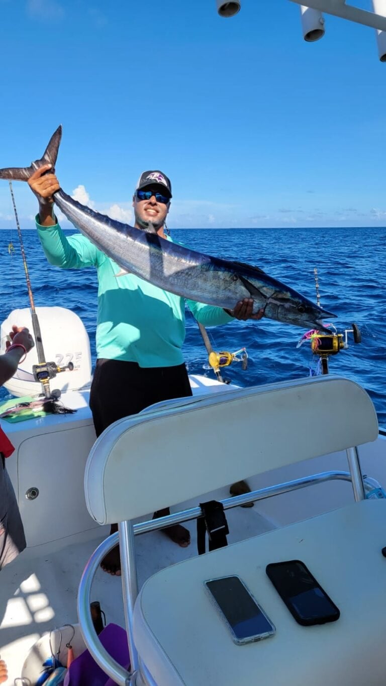 A man in sunglasses holding a large fish on a boat in the clear blue waters off Roatan, Honduras, with fishing gear and the open ocean visible in the background.