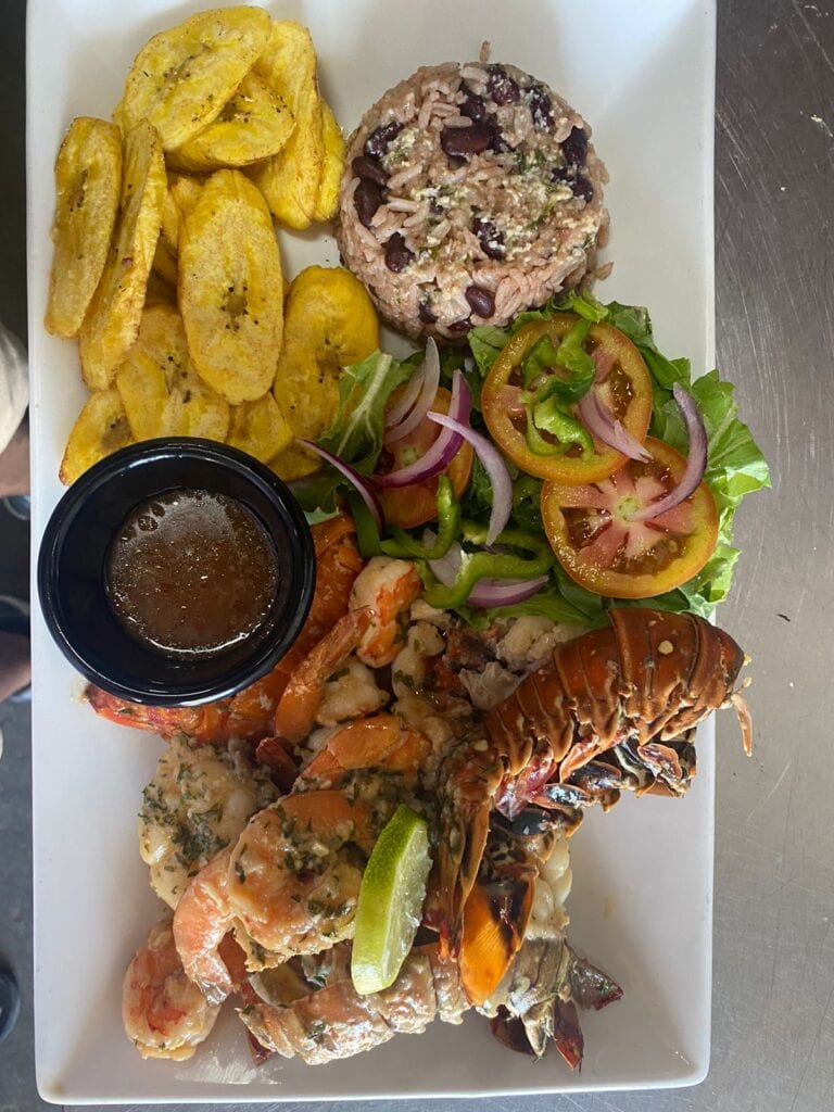 A seafood platter with grilled shrimp, lobster tail, sliced tomatoes, onions, and bell peppers, served with a side of rice and plantains in a restaurant setting on Roatan, Honduras.