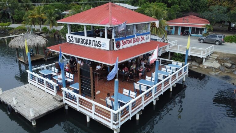 A two-story waterfront restaurant in Roatan, Honduras, featuring outdoor seating on a wooden deck with views of the water, palm trees, and tropical surroundings.
