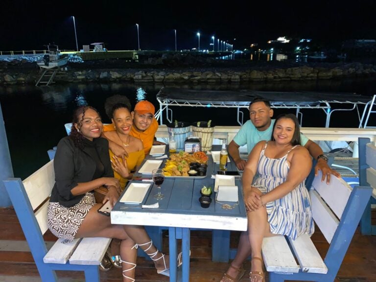 A group of five people enjoying a dinner at a waterfront restaurant in Roatan, Honduras, with a night view of the harbor, boats, and illuminated pier in the background.