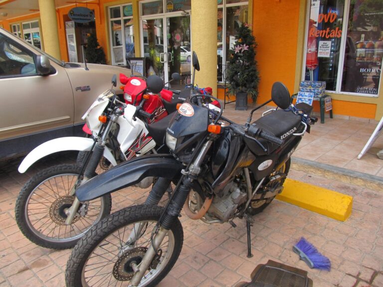 Multiple motorcycles and a beige pickup truck are parked outside a colorful business storefront in Roatan, Honduras, with signage indicating a rental service and the entrance decorated with plants and promotional posters.
