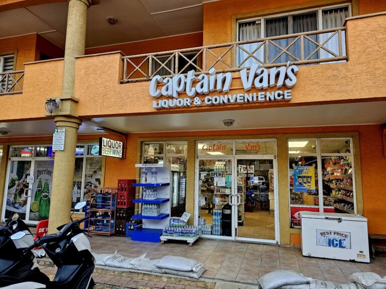 A convenience store on Roatan, Honduras, features an orange exterior with a "Captain Van's Liquor & Convenience" sign above the entrance, displaying various beverages and snacks inside through glass doors, with scooters parked outside on the sidewalk.