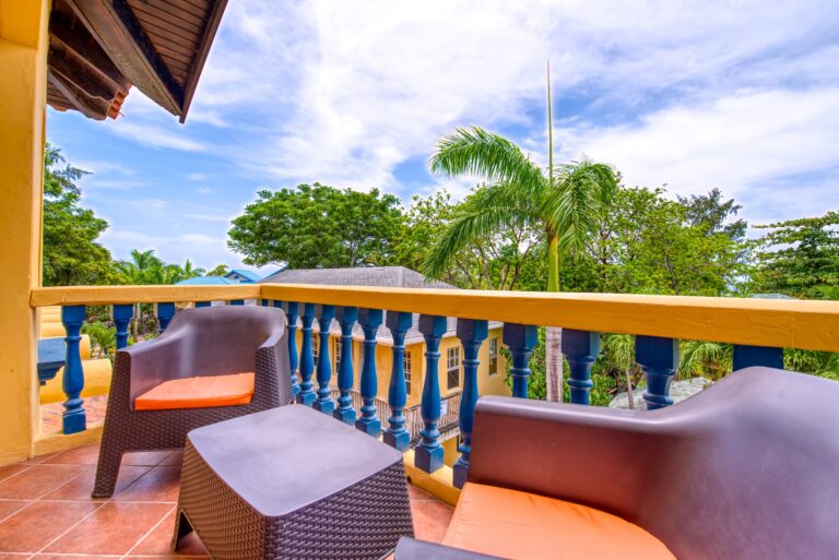 A balcony on a building in Roatan, Honduras, with wicker chairs, a colorful yellow and blue railing, and lush green trees and palm fronds in the background under a partly cloudy sky.