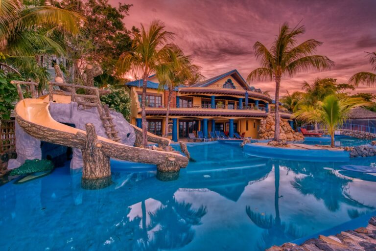 A tropical resort in Roatan, Honduras, features a large multi-story building with blue accents, surrounded by palm trees, a swimming pool with a small rock waterfall, and colorful poolside lounge chairs under a vibrant pink sunset sky.
