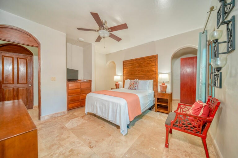 A bedroom in Roatan, Honduras features a large bed with white bedding and orange accents, wooden furniture including a dresser and headboard, a ceiling fan, a television, a red decorative bench, and a window with turquoise curtains.