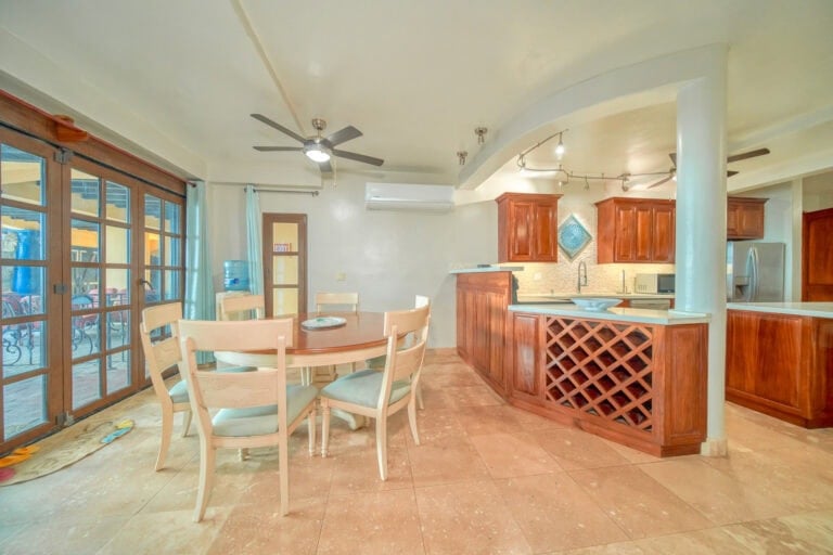 A bright, open kitchen and dining area in a Caribbean-style home on Roatan, Honduras, featuring wooden cabinetry, a round dining table with chairs, sliding glass doors with curtains leading to a balcony, and ceiling fans.