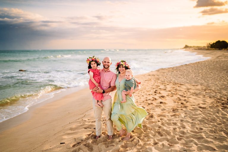 A family of four, wearing flower crowns, enjoying a walk along the sandy beach in Roatan, Honduras at sunset with the ocean and a cloudy sky in the background.