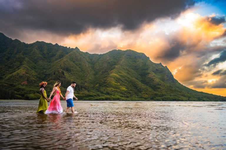 Three people, two women in colorful dresses and a man, walk hand in hand through shallow water near the coast with lush green mountains and a dramatic cloudy sunset sky over Roatan, Honduras.