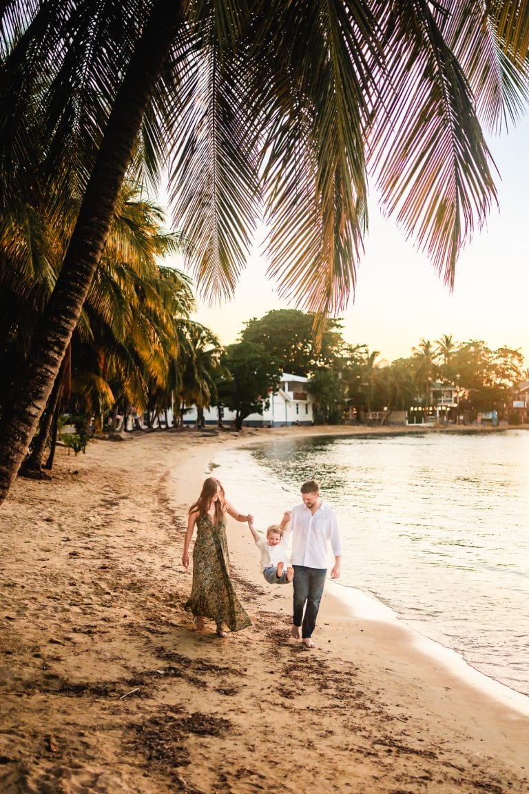A family walks along the sandy beach of Roatan, Honduras at sunset, with palm trees and beachfront houses visible in the background.
