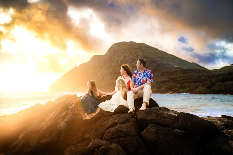 A family sits on rocks by the ocean at sunset on Roatan, Honduras, with a lush mountain in the background and a colorful sky filled with clouds.