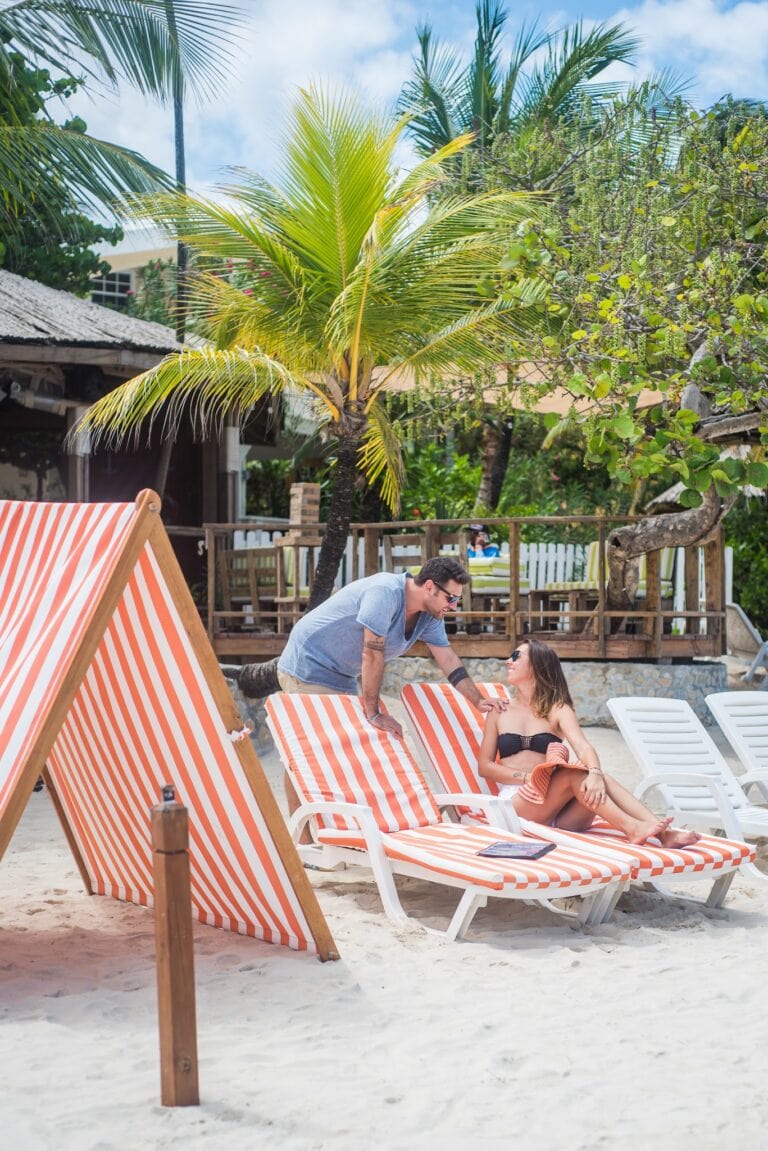 People relaxing and socializing on striped orange and white lounge chairs at a beach in Roatan, Honduras, with palm trees, a wooden fence, and beachside structures visible in the background.