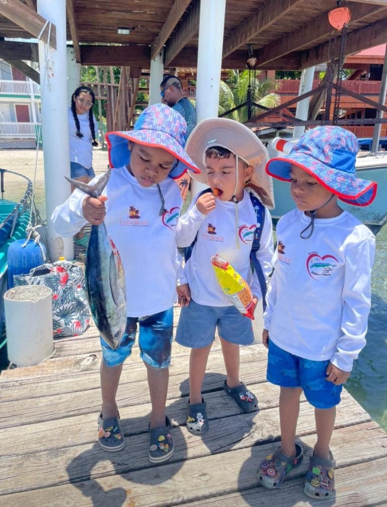 Three young children wearing sun hats and white shirts with the Little Beach Resort logo, stand on a wooden dock in Roatan, Honduras, with one child holding a fish and the others eating snacks, while adults and a boat are visible in the background.