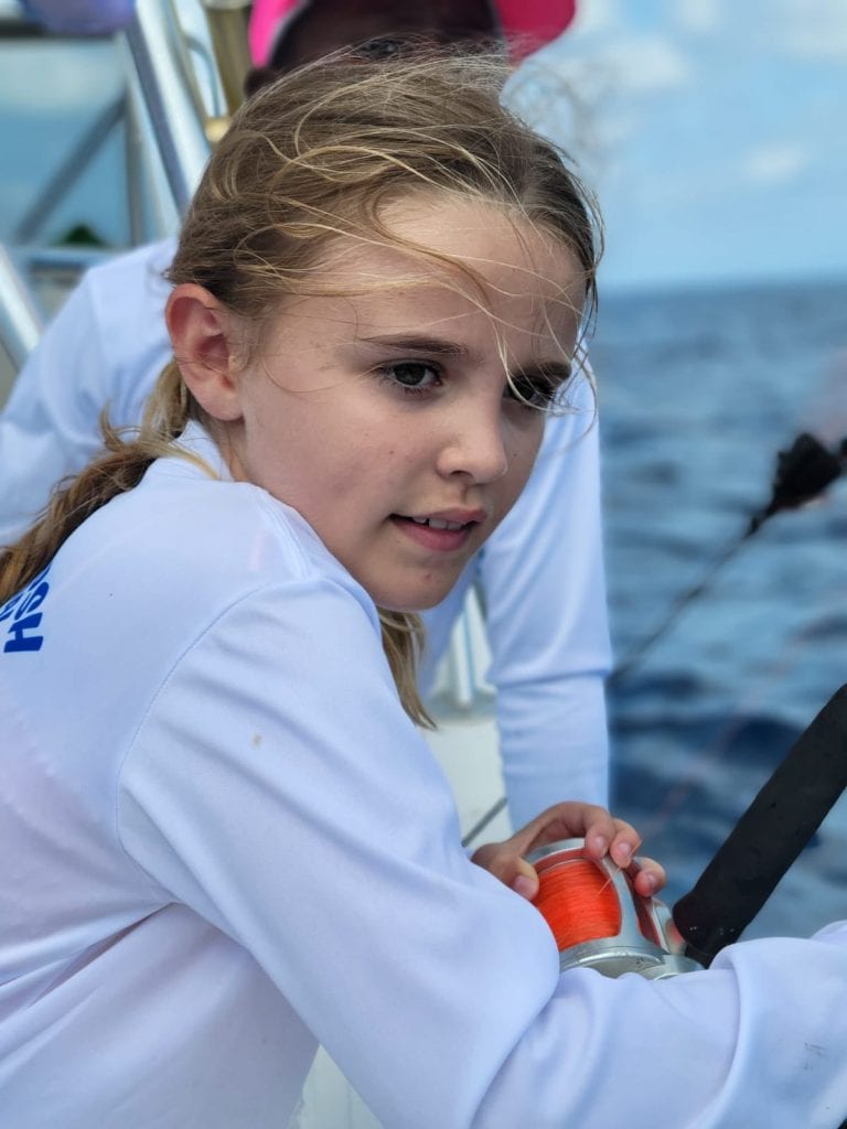A young woman with blonde hair and a white shirt holds a fishing reel aboard a boat in the blue waters near Roatan, Honduras.