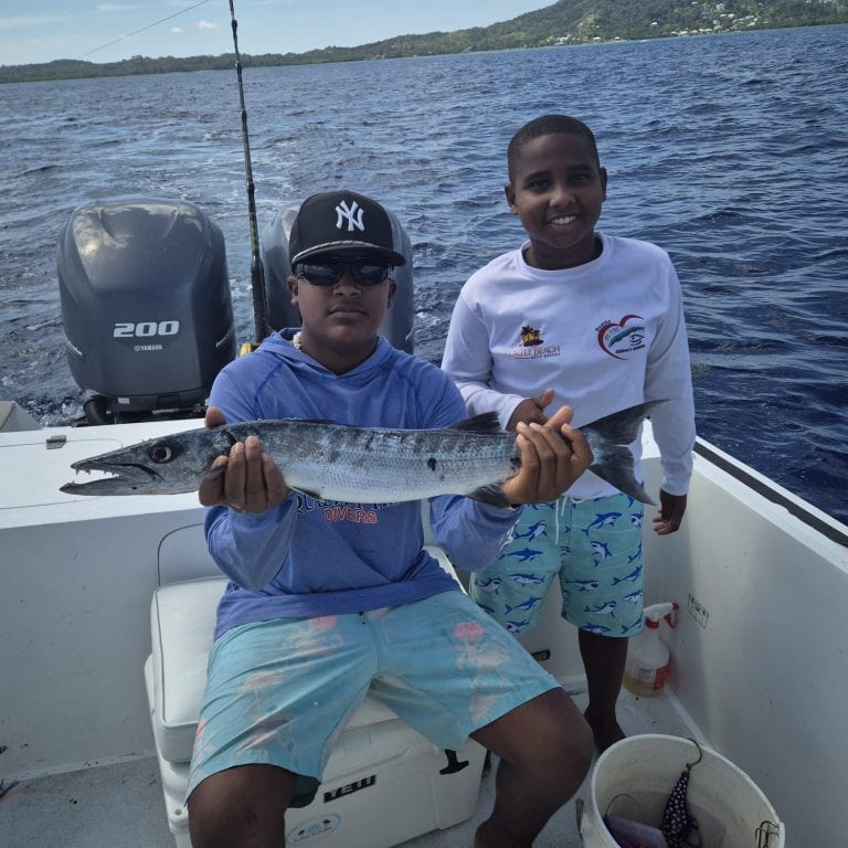 Two boys on a boat in Roatan, Honduras, display a large fish caught during a fishing trip, with ocean water, distant shoreline, and lush green hills in the background.