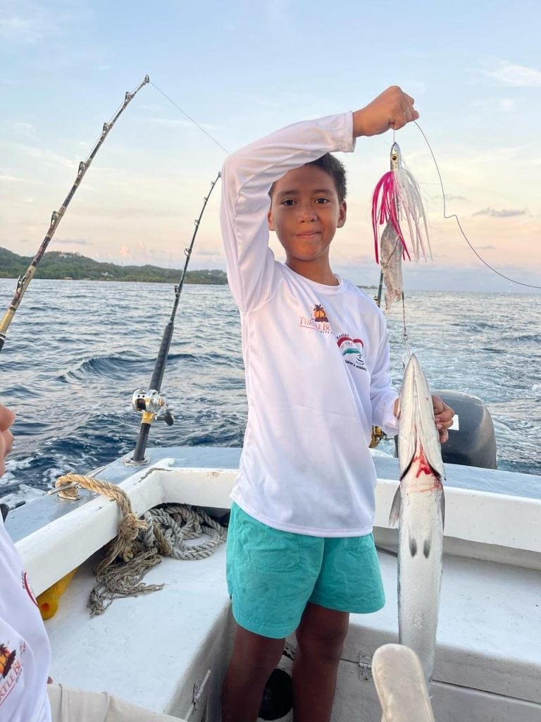 A young boy on a boat in Roatan, Honduras, displays a freshly caught fish with fishing gear and the ocean in the background during a sunset.