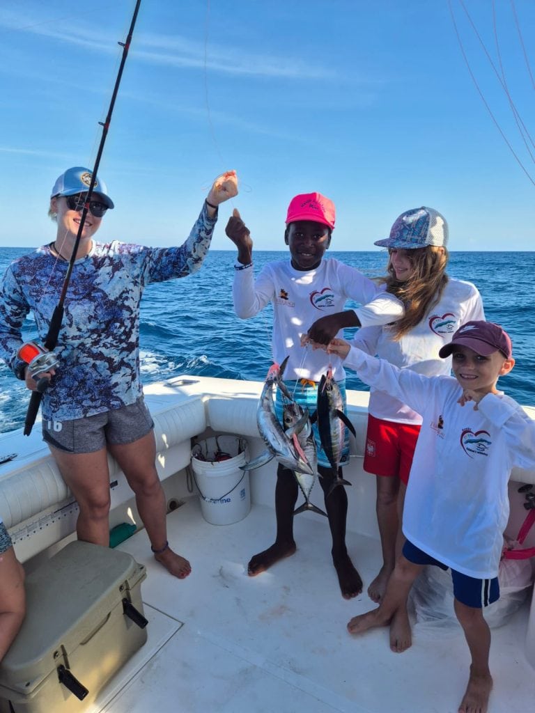 A group of children and a woman are on a boat in Roatan, Honduras, holding freshly caught fish while enjoying a sunny day on the Caribbean Sea.