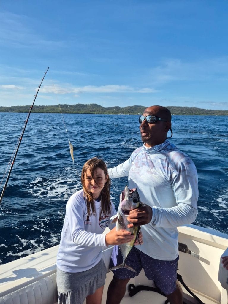 A man and a girl on a boat in Roatan, Honduras, are holding a large fish they caught during a fishing trip against a backdrop of the open sea, distant green hills, and a blue sky.
