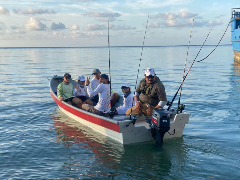A group of people on a small fishing boat with fishing rods in the calm waters of Roatan, Honduras, under a partly cloudy sky.