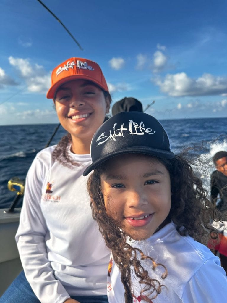 Two smiling girls wearing "Salt Life" hats and white shirts are on a boat in the ocean near Roatan, Honduras, with the blue sky and scattered clouds in the background.