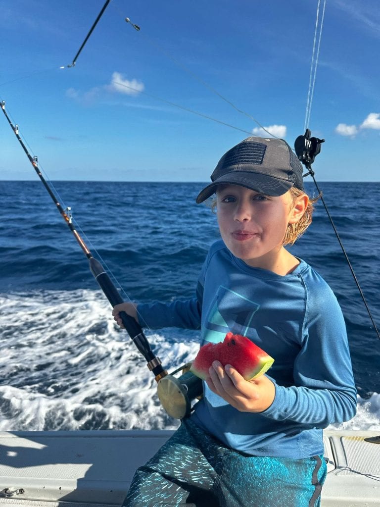 A young boy with a baseball cap fishing on a boat in the blue waters of Roatan, Honduras, while holding a slice of watermelon and smiling against a clear sky.