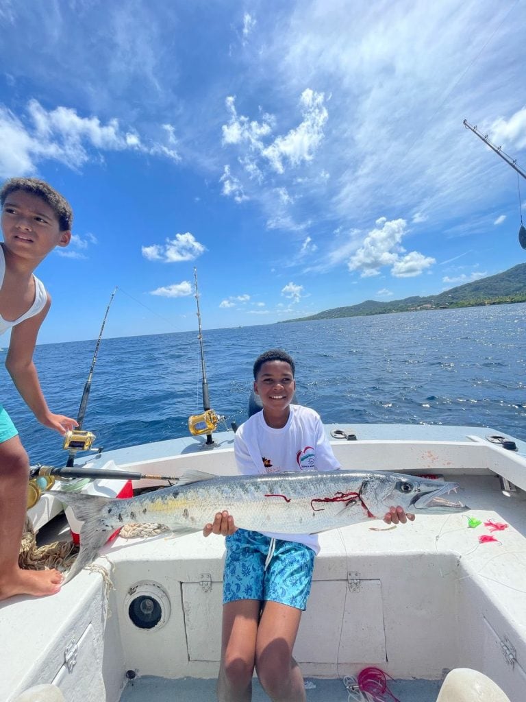 Two boys are on a boat in Roatan, Honduras, holding a large fish they caught, with fishing poles and the ocean visible in the background under a partly cloudy sky.