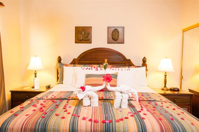 A decorated hotel bedroom in Roatan, Honduras features a large bed with a striped bedspread, floral towel swans with a flower on their necks, rose petal hearts, and two towel animals, illuminated by two bedside lamps and framed by wall art of leaf motifs.