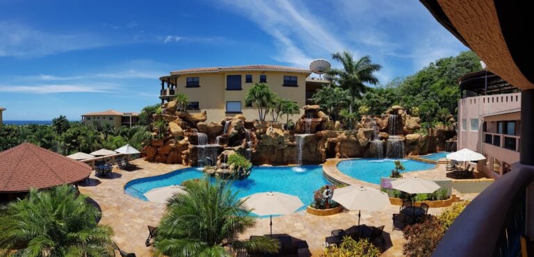 A luxurious hotel pool area in Roatan, Honduras, featuring multiple waterfalls cascading over rocky formations, surrounded by palm trees, lounge chairs with umbrellas, and multi-story buildings with terraces and windows under a bright blue sky.