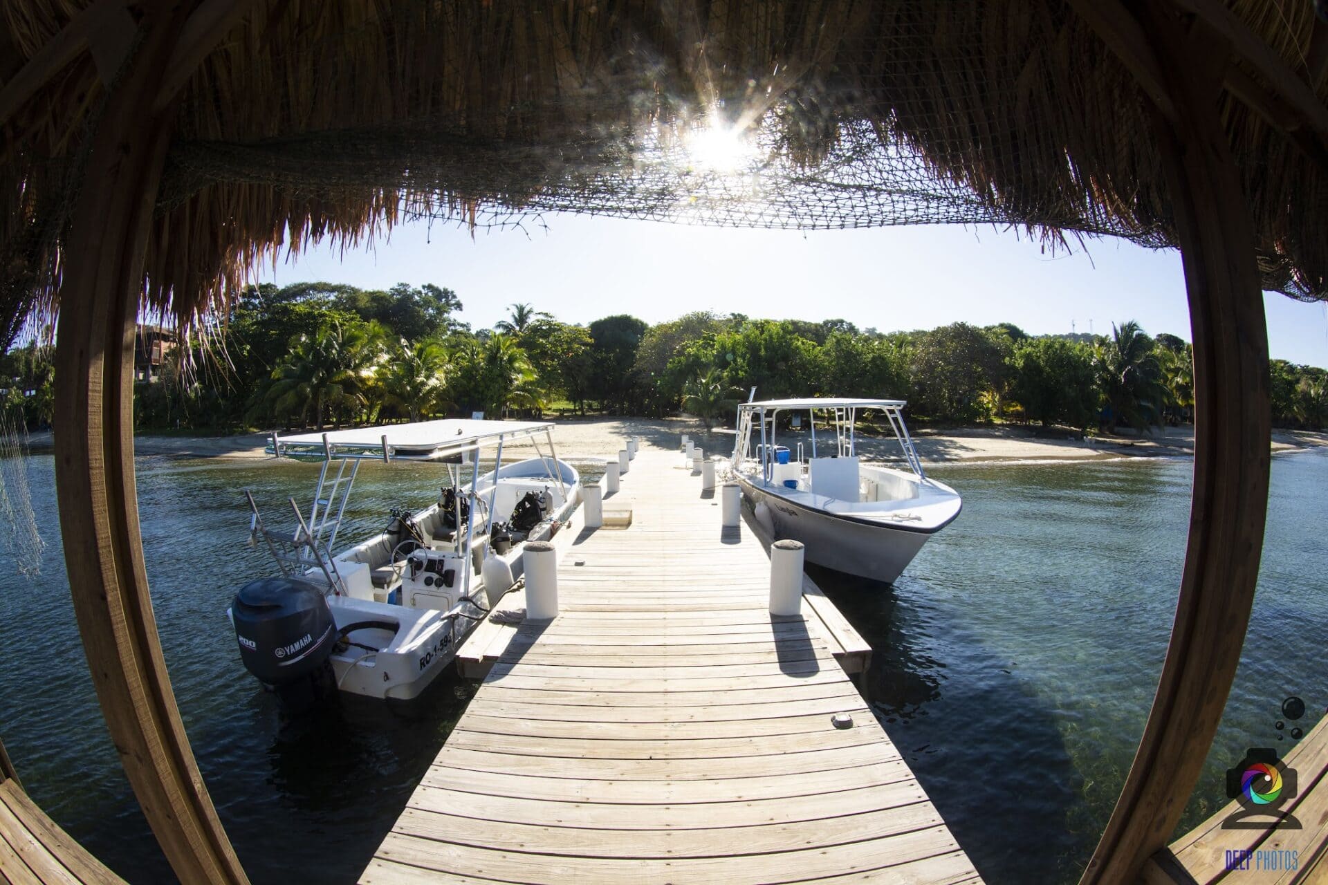 A wooden dock with boats moored, viewed from a thatched-roof hut over calm water surrounded by lush green trees on the coast of Roatan, Honduras.