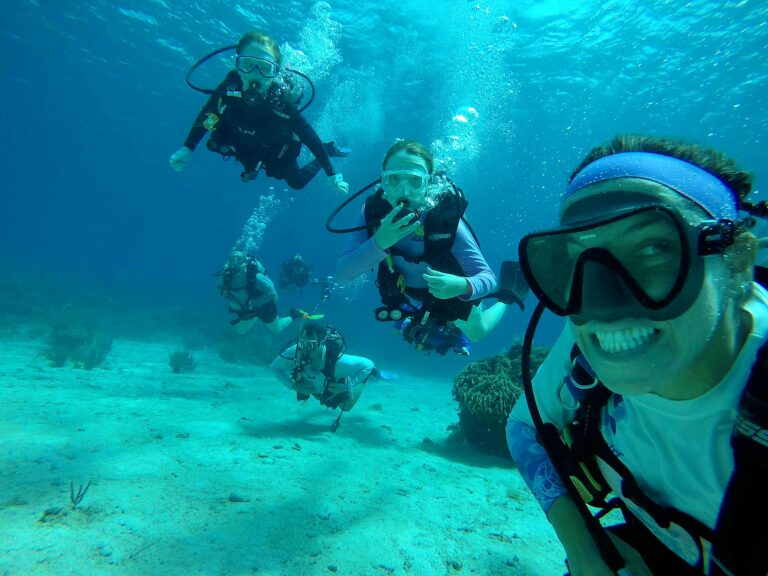 Multiple scuba divers wearing masks and wetsuits swim underwater near coral formations in Roatan, Honduras.