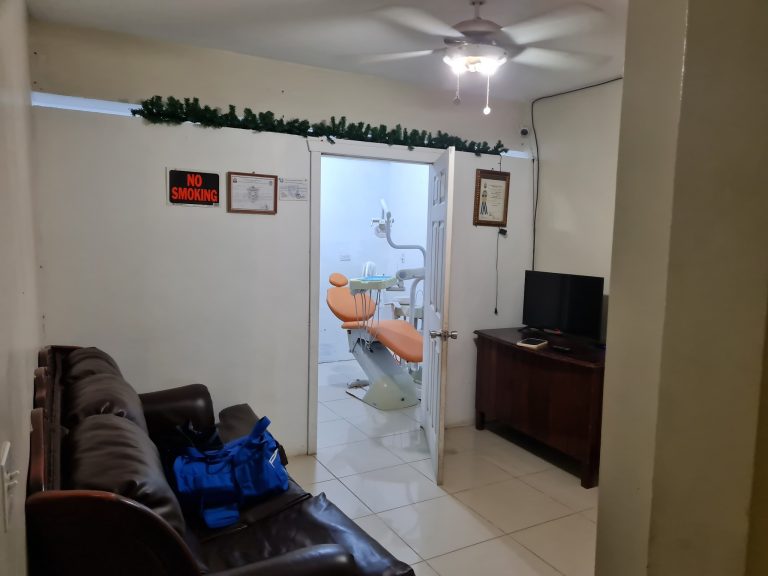 A dental office in Roatan, Honduras, featuring a dental chair with an orange cushion visible through an open doorway, surrounded by plain white walls and framed certificates, with a "No Smoking" sign and decorative greenery above the doorway.