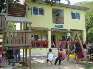 A yellow two-story building in Roatan, Honduras, serves as a community gathering spot with children and adults playing on a wooden playground and swing set outside, while some people are seated on the porch and others are standing nearby.