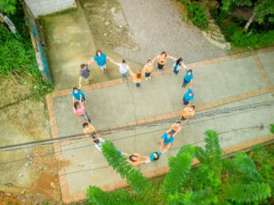 A group of people wearing masks and colorful shirts form a heart shape while holding hands on a concrete pathway surrounded by greenery in Roatan, Honduras.