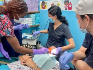 A team of dental professionals wearing face masks and gloves providing dental care to a patient in a clinic with colorful wall art and shelves stocked with supplies in Roatan, Honduras.