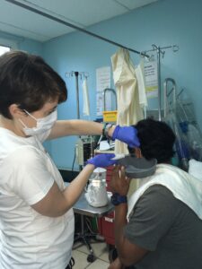 A healthcare worker in a white uniform and face mask administers a medical injection to a patient in a clinical room on Roatan, Honduras.
