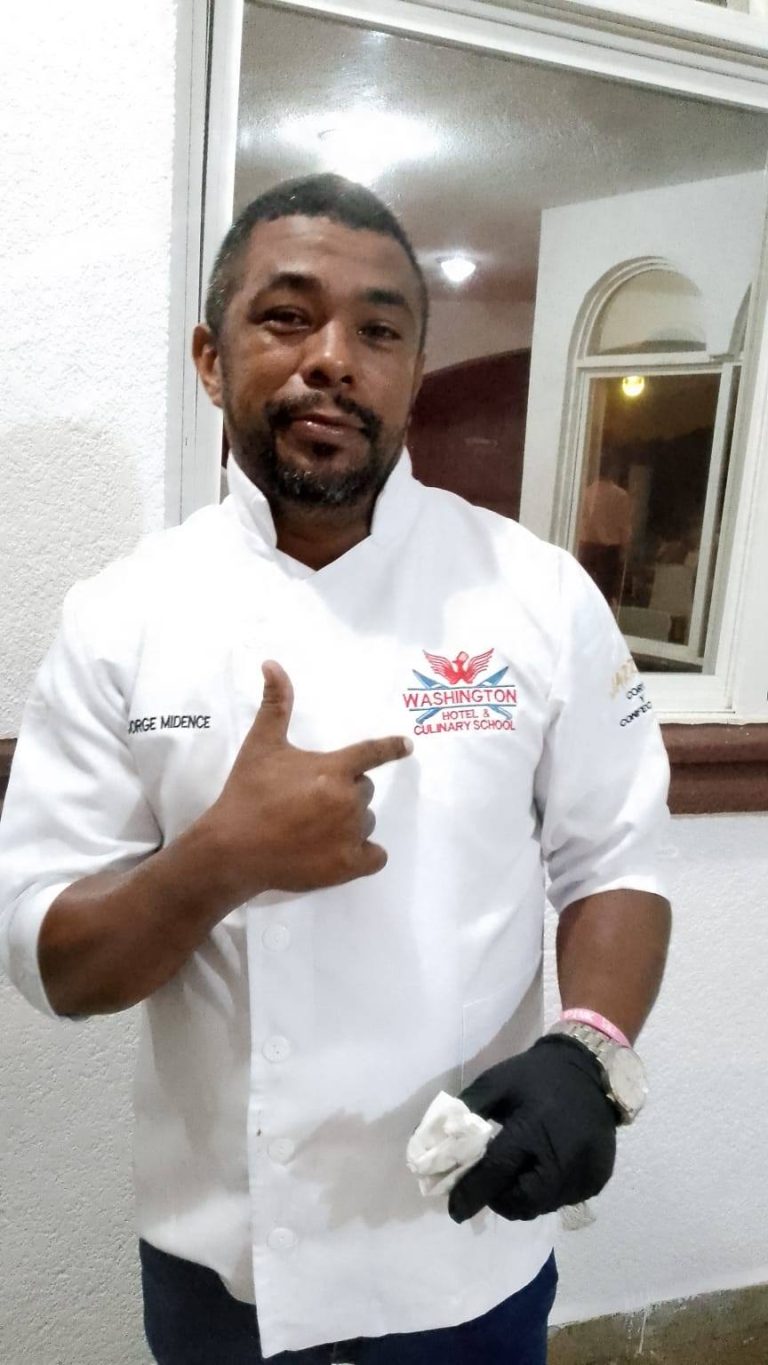 A man wearing a white chef's jacket with "Washington Hotel & Culinary School" and "Jorge Midence" embroidered on it is pointing to the logo on his chest and standing indoors in Roatan, Honduras.