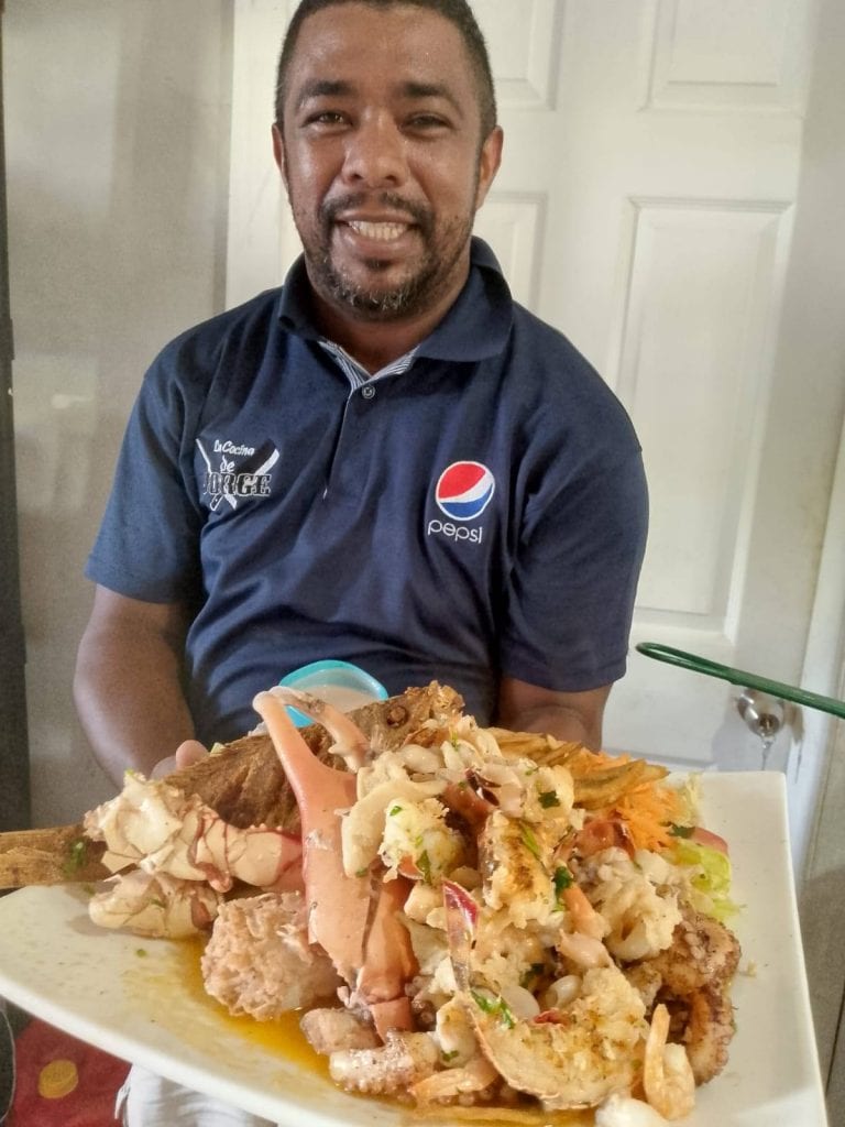 A man wearing a Navy Pepsi shirt holds a large plate of traditional seafood and lobster dishes in a home setting in Roatan, Honduras.