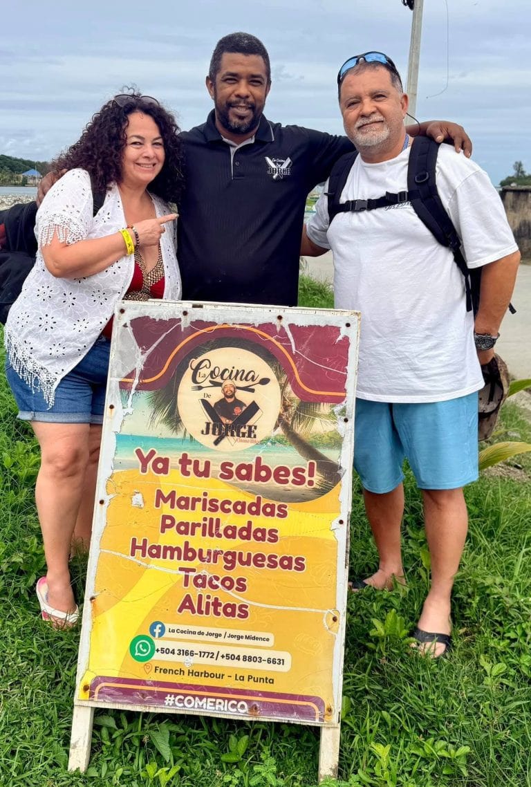 Three people standing outdoors in Roatan, Honduras, posing next to a colorful restaurant sign with menu items in Spanish, with a cloudy sky and greenery in the background.