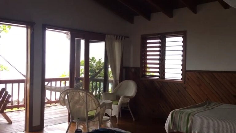 A cozy interior of a beachfront cabana in Roatan, Honduras, featuring a wooden deck visible through large windows and sliding glass doors, with wicker chairs and a bed with striped bedding, all surrounded by natural wood accents and views of lush greenery outside.