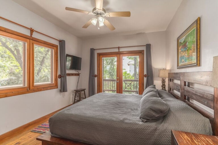 A cozy bedroom in a Roatan, Honduras vacation rental features wooden-framed windows and doors, a ceiling fan, a wall-mounted flat-screen TV, a painting of local scenery, and a balcony overlooking lush tropical greenery.