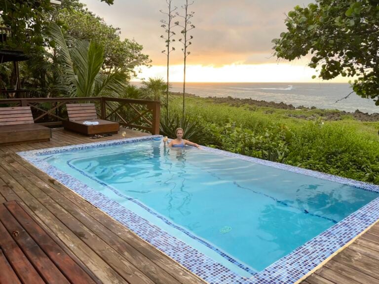 A woman relaxes in a swimming pool overlooking lush greenery and the ocean at sunset, with a boat passing in the distance on Roatan, Honduras.