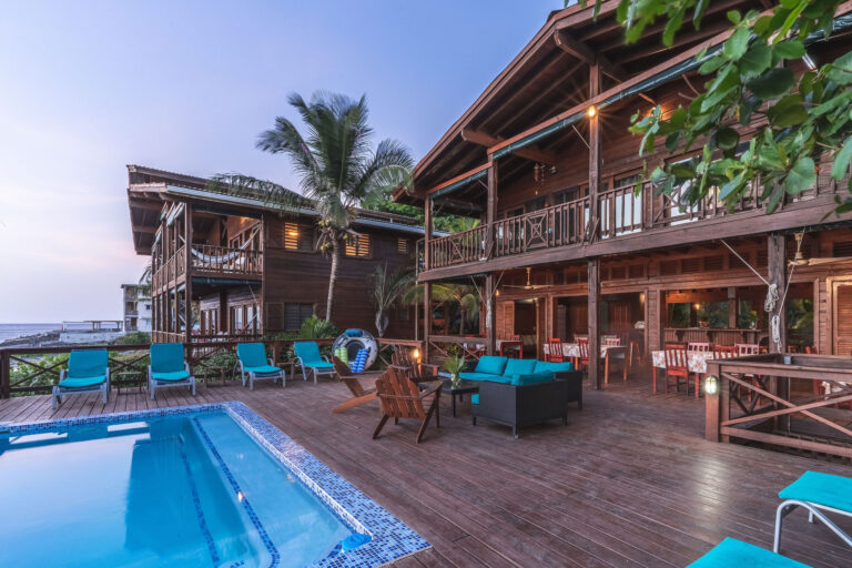 A wooden deck area with outdoor seating, lounge chairs, and a swimming pool overlooking the ocean in Roatan, Honduras, with a two-story wooden building, lush green plants, and palm trees in the background.