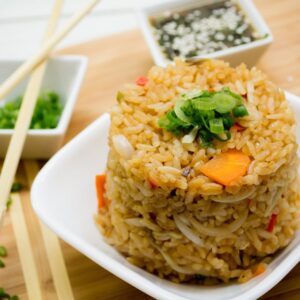 A serving of fried rice topped with chopped green onions is presented on a white plate, accompanied by small bowls of soy sauce and chopped green onions, on a wooden surface in Roatan, Honduras.