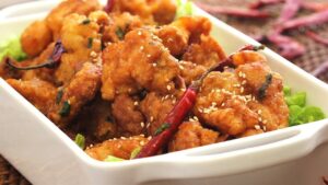 A white rectangular dish filled with spicy fried chicken garnished with sesame seeds, chopped green onions, and a red chili pepper, set on a table in Roatan, Honduras.
