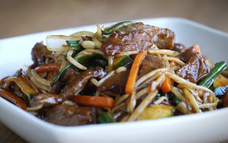 A bowl of stir-fried noodles with vegetables and tender meat, featuring carrots, onions, green beans, and bean sprouts, served in a white dish, representing local cuisine in Roatan, Honduras.